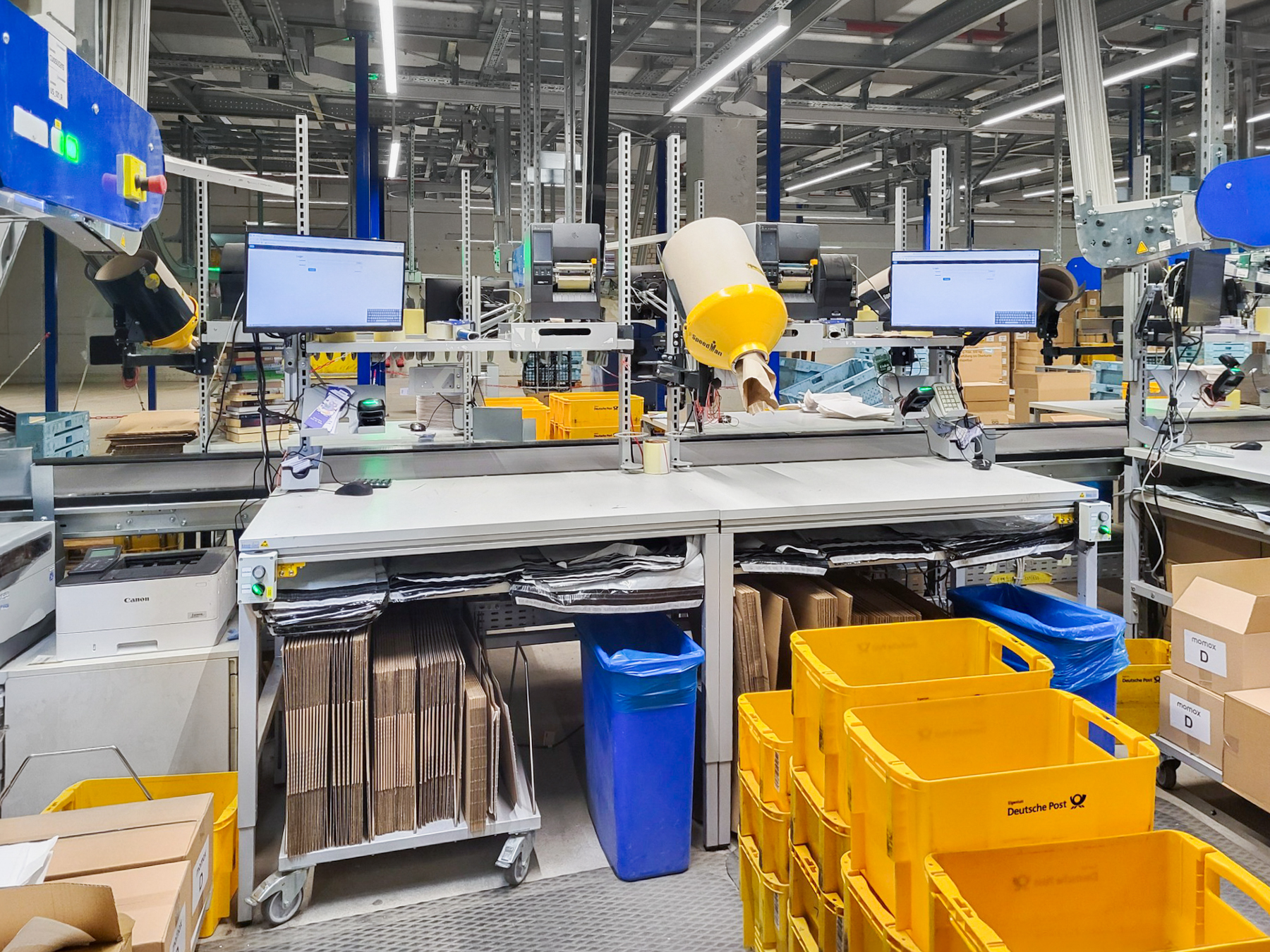 ERGO-line Packtisch im Logistikbereich mit Monitoren, Scanner, Ablageboden, fahrbares Faltschachtelmagazin und Regalen